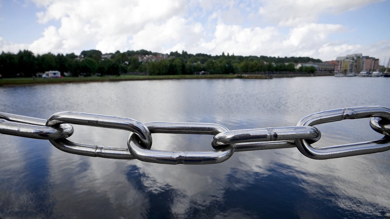 A close-up view of a steel chain link fence overlooking a tranquil lake under a partly cloudy sky.